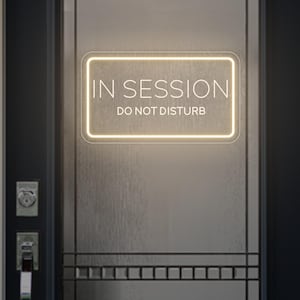Può includere: Un'insegna al neon rettangolare con le parole "IN SESSION" sopra "DO NOT DISTURB" in bianco. L'insegna è delineata da una luce calda e brillante ed è montata su una porta scura con pannelli di vetro. La porta ha una maniglia argentata e una serratura.