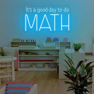 May include: A bright blue neon sign that reads "it's a good day to do MATH" in a classroom setting. The sign is mounted on a light blue wall. The room has wooden shelves, tables, and a potted plant.
