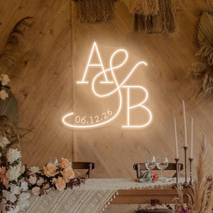 May include: A neon sign with the letters "A & B" and the date "06.12.26" in a cursive font. The sign is lit with a warm, soft glow and is mounted on a wooden wall. The scene is decorated with flowers and candles, suggesting a wedding or event.
