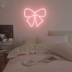 Op de afbeelding: Een roze neon strik-bord hangt boven een bed met roze en groene beddengoed. De kamer heeft een zachte, warme uitstraling. Een kleine tafel met een laptop en andere items is zichtbaar in de hoek.