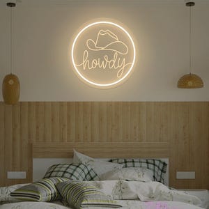 May include: A neon sign with a cowboy hat and the word "howdy" in a circular design. The sign is illuminated with a warm, inviting glow. The sign is mounted on a wall above a bed with pillows and a patterned comforter.