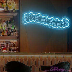 Puede incluir: Un letrero de neón con el texto "it's shoto'clock" en color azul brillante. El letrero está montado en una pared de madera sobre una barra con botellas de licor. Hay dos sillas negras frente a la barra.
