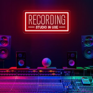 May include: A rectangular red neon sign mounted on a dark wall with the white text RECORDING STUDIO IN USE enclosed in a border. The sign is positioned above a professional audio mixing console with multiple faders, knobs, and studio speakers.