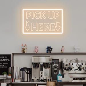 Op de afbeelding: Een rechthoekig neonbord met een warmwitte gloed, gemonteerd aan een muur. Het bord bevat de tekst PICK UP HERE in een vet, schreefloos lettertype, met twee naar beneden wijzende pijlen onder de tekst, aan weerszijden van het woord HERE.
