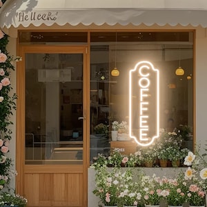 May include: A vertical rectangular neon sign displaying the word COFFEE in glowing white light, mounted on a glass window of a storefront. The shop features a wooden door, a beige awning, and numerous potted plants with pink and white flowers.