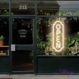 以下が含まれることがあります： 長方形のネオンサイン「OPEN」が光るお店の正面。暗い緑色のドアと窓枠にネオンサインが映えます。ドアの上には数字の「213」があります。お店の内外には緑豊かな植物が見えます。