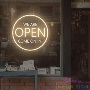 May include: A neon sign in a shop window reads "WE ARE OPEN COME ON IN!" in a circular design. The sign is illuminated with a warm, inviting glow, set against a blurred background of the shop's interior and exterior.