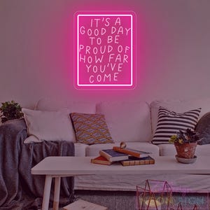 May include: A bright pink neon sign with the text "IT'S A GOOD DAY TO BE PROUD OF HOW FAR YOU'VE COME" hangs above a white couch. The sign is rectangular with a white border. The room has a modern, minimalist aesthetic.