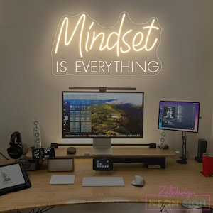 Puede incluir: Un letrero de neón con la frase "Mindset is Everything" en fuente cursiva, montado sobre un escritorio con ordenador, tableta y otros accesorios tecnológicos. El letrero emite un brillo blanco cálido.
