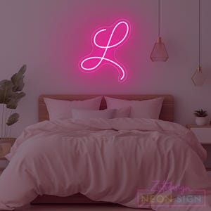 May include: A pink neon sign in the shape of the letter "L" is mounted on a wall above a bed with pink bedding. The room is decorated in shades of pink, with hanging pendant lights and a small plant on a nightstand.