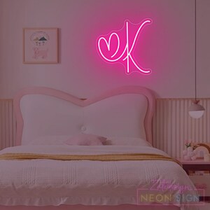 May include: A neon pink sign in the shape of the letter "K" with a heart, mounted on a wall above a bed. The bedroom has a pink and white color scheme, with a bed, a nightstand, and a framed picture.