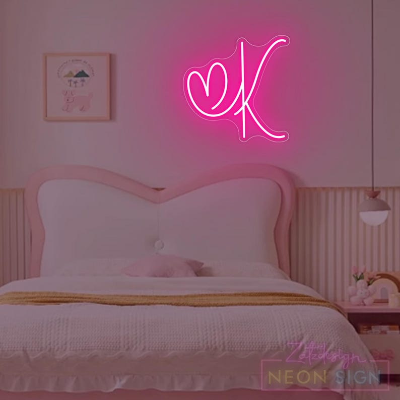 May include: A neon pink sign in the shape of the letter "K" with a heart, mounted on a wall above a bed. The bedroom has a pink and white color scheme, with a bed, a nightstand, and a framed picture.