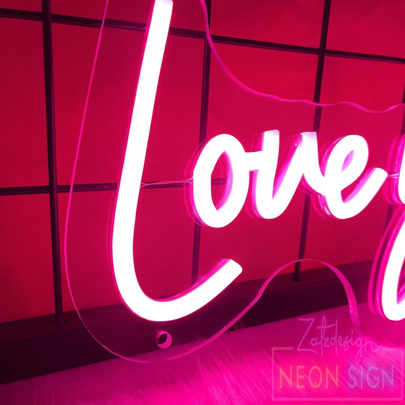 May include: A neon sign in a cursive font that reads "love you always" in bright white against a pink background. The sign is mounted on a clear acrylic backing. The Zatzdesign Neon Sign logo is visible.