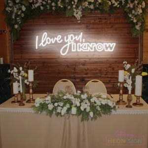 May include: A wedding table setting with a neon sign saying "I love you, I know". The table is decorated with white flowers, candles, and a beige tablecloth. The backdrop is made of wooden panels.