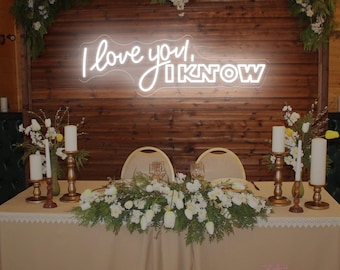 May include: A wedding table setting with a neon sign saying "I love you, I know". The table is decorated with white flowers, candles, and a beige tablecloth. The backdrop is made of wooden panels.