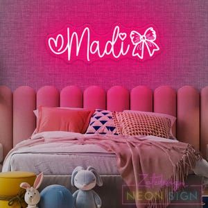 May include: A neon sign in the shape of the name "Madi" in a cursive font, with a heart and bow detail. The sign is bright pink and mounted on a pink textured wall above a bed with pink bedding and decorative pillows.