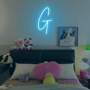 May include: A neon sign in the shape of the letter "G" glows brightly on a wall. Below, a bed is adorned with plush toys and pillows, including a large pink cloud-shaped pillow. A painting of a cat hangs on the wall.