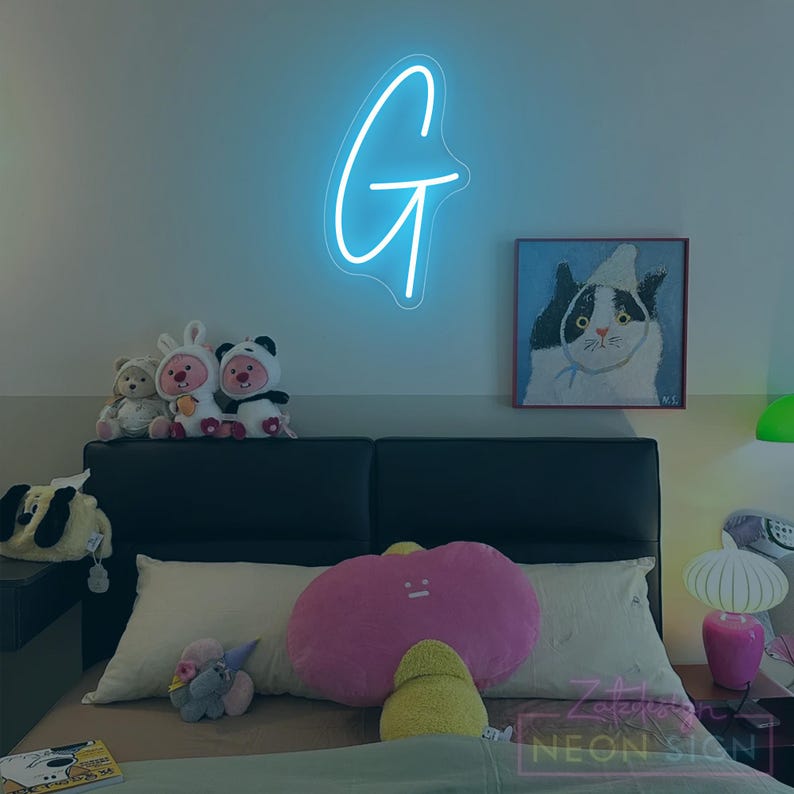 May include: A neon sign in the shape of the letter "G" glows brightly on a wall. Below, a bed is adorned with plush toys and pillows, including a large pink cloud-shaped pillow. A painting of a cat hangs on the wall.
