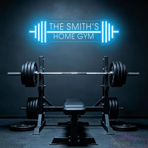 May include: A bright blue neon sign mounted on a dark wall above a weight bench and barbell set. The sign is shaped like a barbell and features the text THE SMITH'S HOME GYM in a clean, sans-serif font, glowing against the dark background.