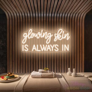 May include: A wall-mounted neon sign with white glowing script and block lettering that reads glowing skin IS ALWAYS IN. The sign is positioned above a massage table featuring a folded beige towel, a small bowl, and two white herbal balls.