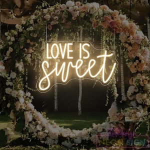 May include: A neon sign with the words "LOVE IS sweet" in a cursive font, hanging in front of a floral arch. The sign glows with a warm, inviting light, and the flowers are a mix of white, pink, and peach.