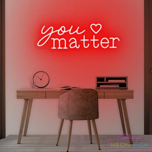 May include: A red neon sign with the words "you matter" in a cursive font, with a heart symbol. The sign is mounted on a wall above a wooden desk with a chair. A clock and desk organizer are on the desk.