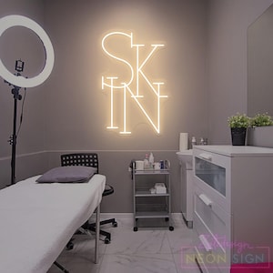 May include: A neon sign displaying the word "SKIN" in a beauty salon setting. The sign glows with a warm white light against a grey wall. The room includes a treatment bed, a trolley, and a ring light, creating a professional atmosphere.