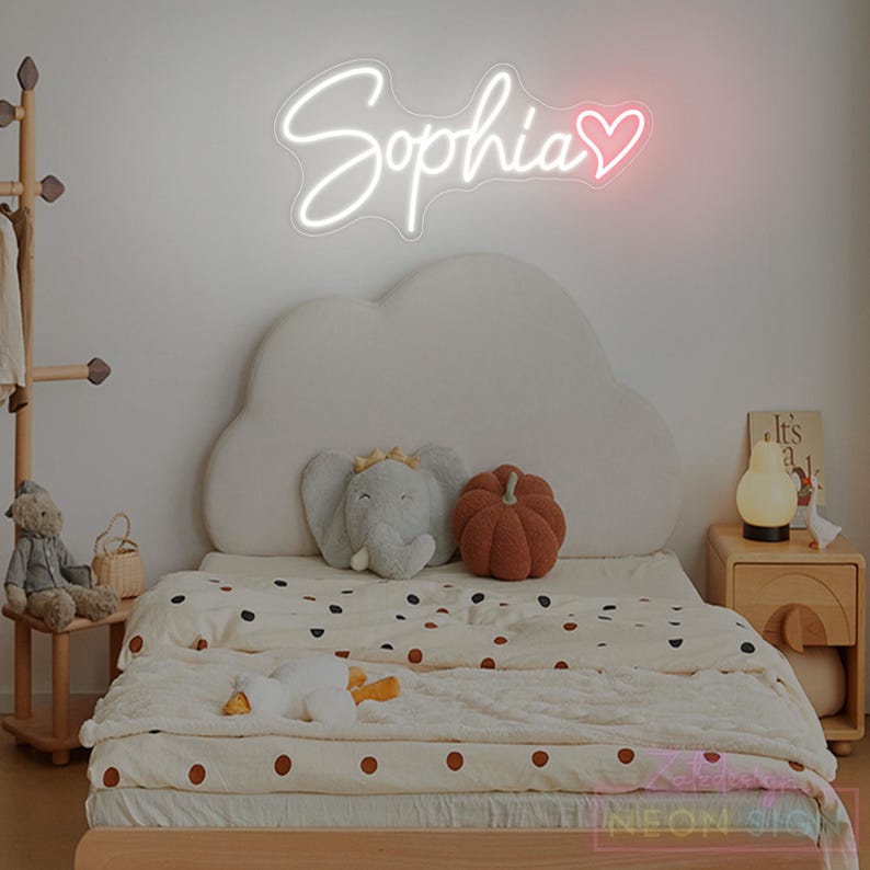 May include: A neon sign displaying the name "Sophia" in white cursive script with a pink heart. The sign is above a bed with a cloud-shaped headboard, a polka-dot blanket, and decorative pillows. A wooden bedside table and coat rack are also in view.