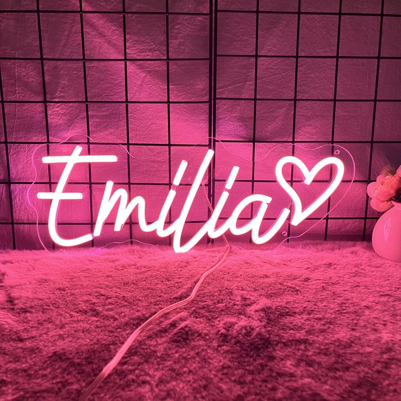 May include: A pink neon sign displaying the name "Emilia" in a cursive script, finished with a heart shape. The sign is lit up against a pink backdrop, with a grid pattern visible behind it.
