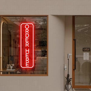 May include: A vertical rectangular neon sign with a bright red glow displaying the text ORDER HERE in a clean sans-serif font. The sign is mounted inside a glass window of a building with a light-colored tiled exterior wall and a wooden door frame.