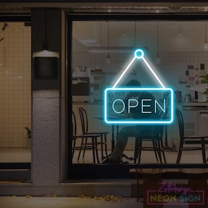 Pode incluir: Uma placa de néon com a palavra "OPEN" em azul brilhante, pendurada numa montra. A placa é delineada por um brilho azul brilhante, com um triângulo branco por cima. O interior da loja é visível através do vidro.