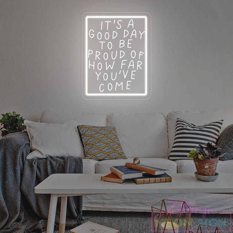 May include: A white neon sign with the text "IT'S A GOOD DAY TO BE PROUD OF HOW FAR YOU'VE COME" hangs on a wall above a white couch. The sign is rectangular with a white border. The room has a coffee table with books and a plant.