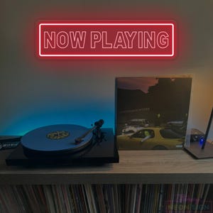 May include: A red neon sign displaying "NOW PLAYING" above a record player with a blue vinyl record. A record album is displayed to the right. The setup is on a wooden shelf with a collection of records below.