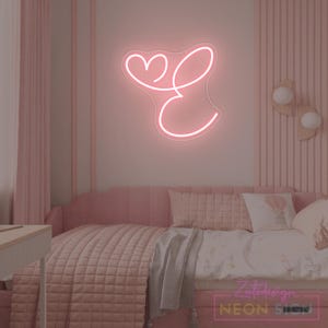 May include: A pink neon sign in the shape of the letter "E" with a heart above it, mounted on a pink wall. The sign is in a bedroom with pink bedding and decor. The sign is from ZeteDesign Neon Sign.