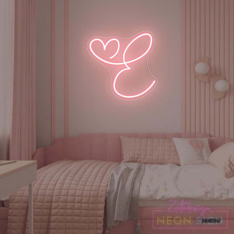 May include: A pink neon sign in the shape of the letter "E" with a heart above it, mounted on a pink wall. The sign is in a bedroom with pink bedding and decor. The sign is from ZeteDesign Neon Sign.