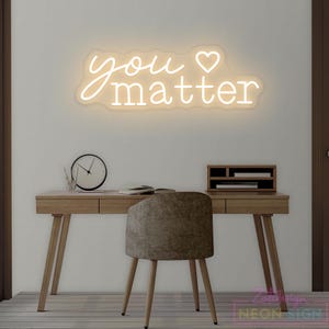 May include: A neon sign with the words "you matter" in a cursive font, with a heart symbol. The sign is mounted on a white wall above a wooden desk with a chair, clock, and desk organizer. The sign is a decorative item.