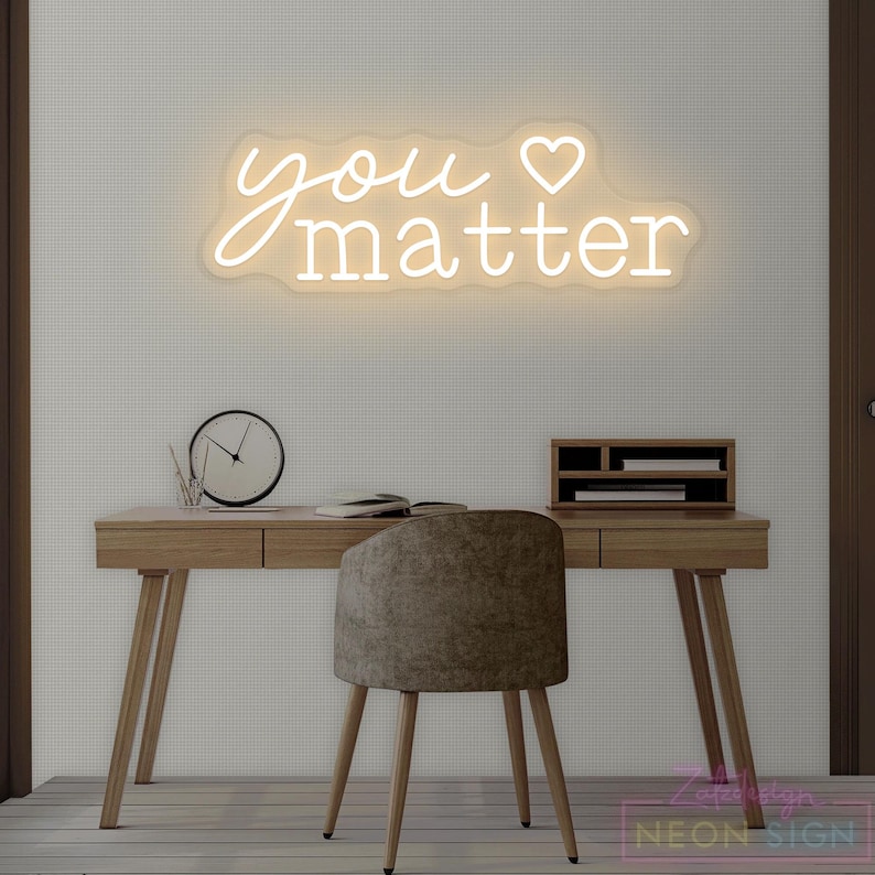 May include: A neon sign with the words "you matter" in a cursive font, with a heart symbol. The sign is mounted on a white wall above a wooden desk with a chair, clock, and desk organizer. The sign is a decorative item.