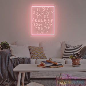 May include: A pink neon sign with the text "IT'S A GOOD DAY TO BE PROUD OF HOW FAR YOU'VE COME" hangs on a wall above a white couch. The sign is rectangular with a white border. A white coffee table holds books and a potted plant.