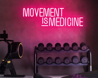 May include: A neon sign with the words "MOVEMENT IS MEDICINE" in bright pink, mounted on a wall. The gym setting includes a rack of dumbbells and exercise equipment, promoting fitness and wellness.