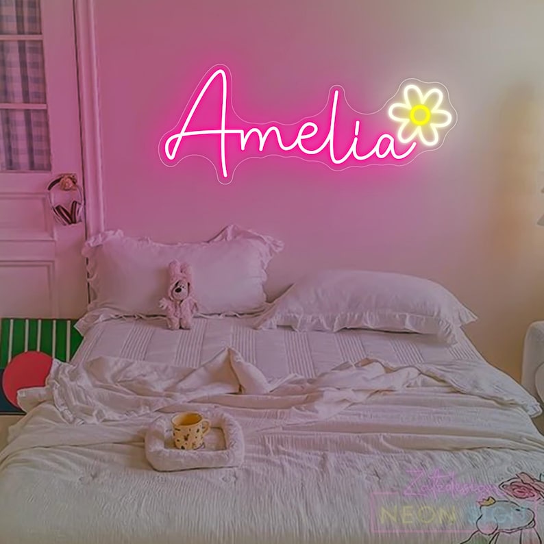 May include: A pink neon sign displaying the name "Amelia" in cursive, accompanied by a daisy design, hangs above a bed. The bed is adorned with white bedding, pillows, and a soft toy. The room has a gentle, inviting atmosphere.