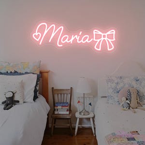 May include: A pink neon sign spelling "Maria" with a heart and bow design, mounted on a white wall above two beds. The sign illuminates a bedroom setting with two beds, a lamp, and small chairs.