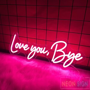 May include: A neon sign with the words "Love you, Bye" in a cursive font. The sign is white and mounted on a clear acrylic backing. The background is a red tiled wall and pink fluffy surface.