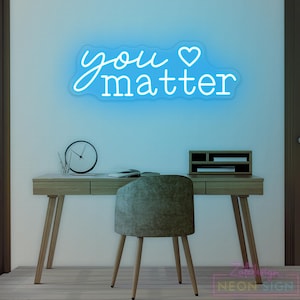 May include: A blue neon sign with the words "you matter" and a heart symbol is mounted on a light blue wall. Below, a wooden desk with a chair, clock, and office supplies.