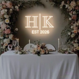 May include: A wedding reception table is set with a white tablecloth, floral arrangements, and candles. Above the table, a neon sign displays the initials "HK" and the year "EST 2026". The background features floral decorations.