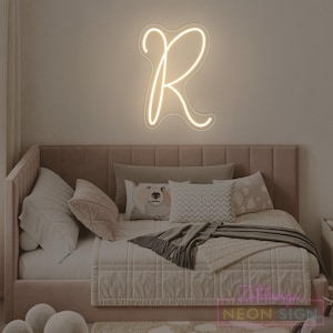 May include: A neon sign in the shape of the letter "R" glows with a soft, warm light. The sign is mounted on a neutral-colored wall above a bed with a pink headboard and various pillows. The bed is made with a white comforter and a gray throw blanket.