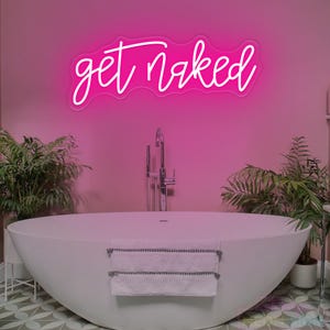May include: A bright pink neon sign featuring the text get naked in a cursive script font. The sign is mounted on a solid pink wall above a white oval bathtub. A white towel with gray stripes hangs over the front edge of the bathtub.