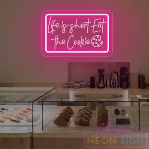 Puede incluir: Un letrero de neón rosa brillante con la frase "Life is short Eat the Cookie" en letra cursiva. Debajo, una vitrina exhibe pasteles y productos horneados.