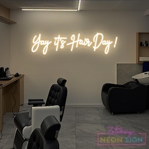 Może przedstawiać: Neonowy szyld w salonie fryzjerskim z napisem "Yay it's Hair Day!" w kursywie. Szyld jest podświetlony ciepłym, białym światłem i zamontowany na neutralnej ścianie. Salon posiada czarne krzesła i umywalkę.