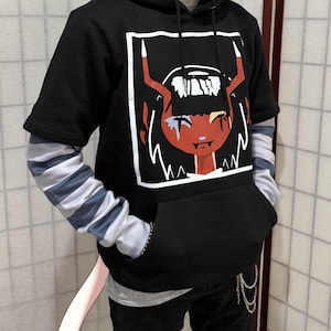 May include: Black hooded sweatshirt with a graphic of a red-skinned character with horns and a white outline. The sweatshirt has a front pocket and is worn over a striped long-sleeve shirt. The sleeves are gray and blue.