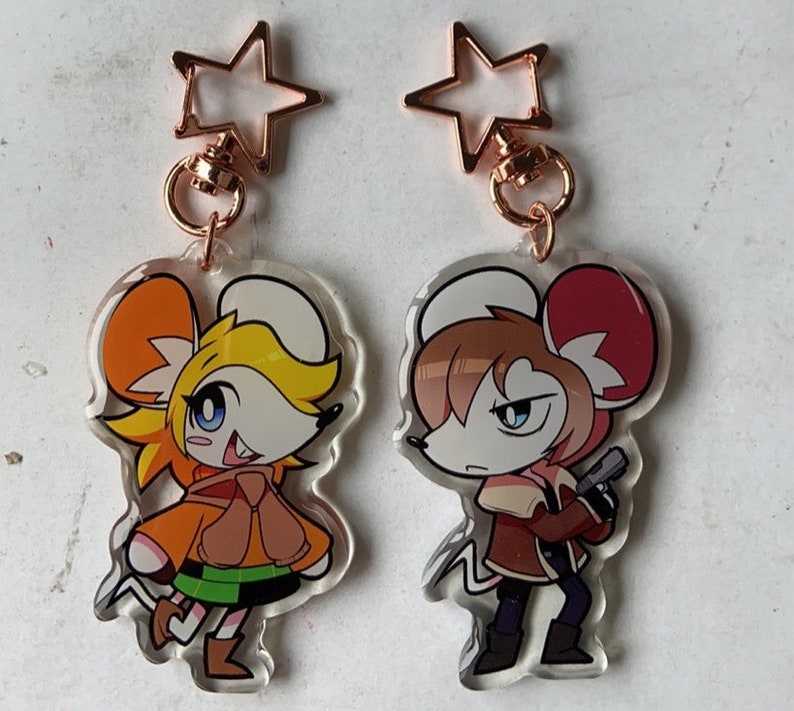 May include: Two cartoon mouse character keychains with gold star charms. The mouse on the left is wearing a brown jacket and green pants. The mouse on the right is wearing a brown jacket and black pants.